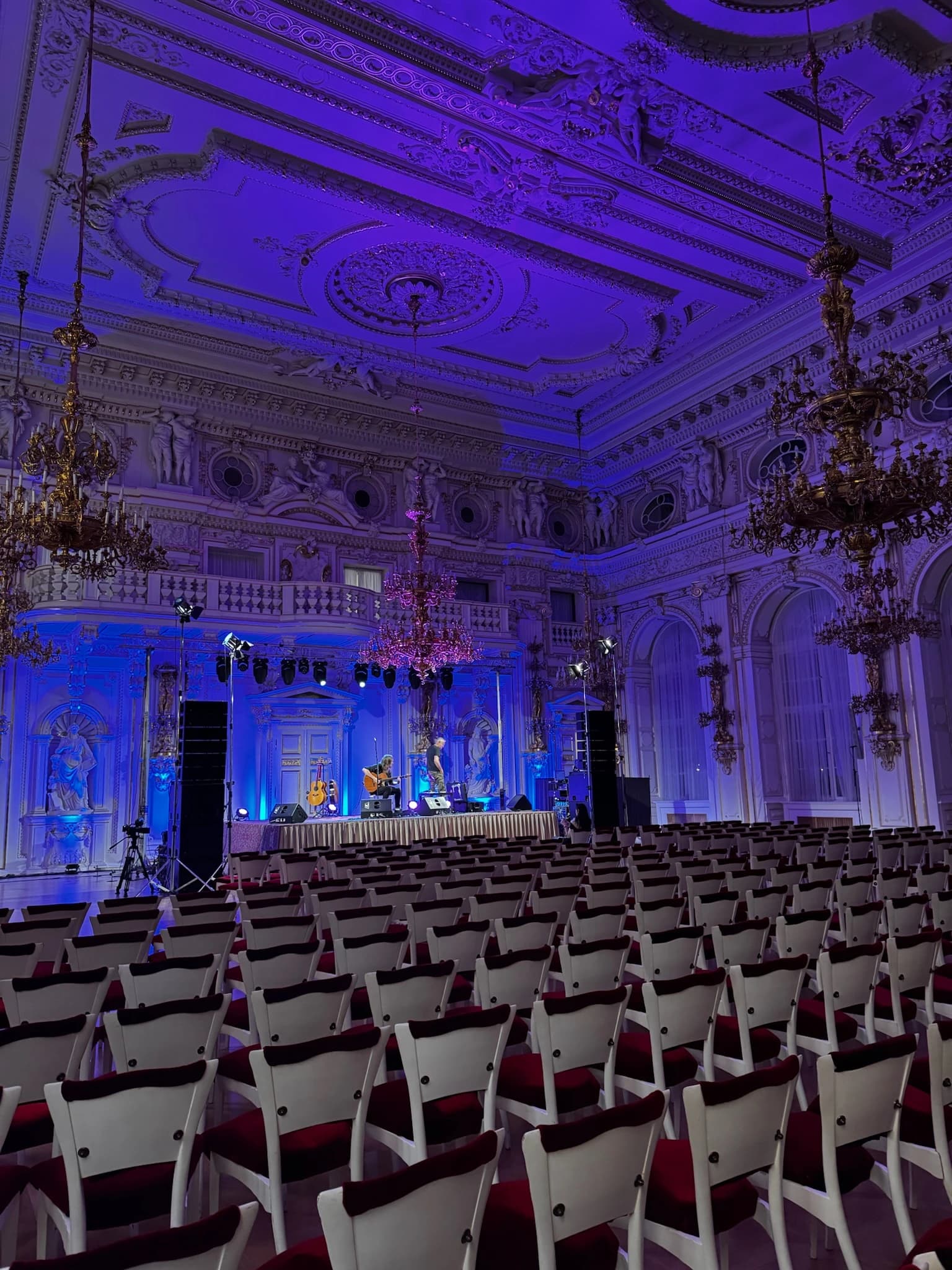 Barokní sál s modrým osvětlením připravený na koncert s řadami židlí